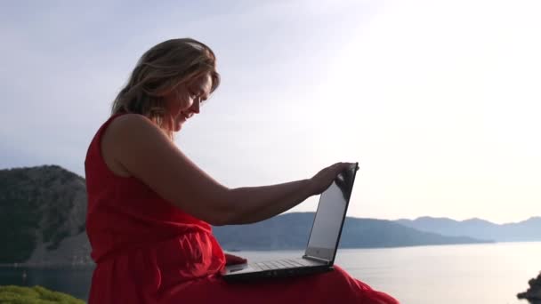 Жінка закінчує роботу над ноутбуком високо на скелі над морем, повільний рух — стокове відео