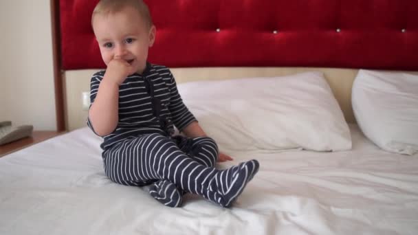 En liten pojke i en randig Jumpsuit hoppar glatt på den stora sängen i ett hotellrum — Stockvideo