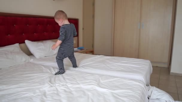 En söt pojke spelar glatt, faller på en stor säng i ett hotellrum, slow motion — Stockvideo