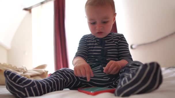 Ein Baby klickt auf den Bildschirm des Smartphones, sitzt auf dem Bett, Zeitlupe — Stockvideo