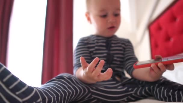 Niño pequeño aplaude alegremente un teléfono inteligente en una cama grande en cámara lenta — Vídeos de Stock