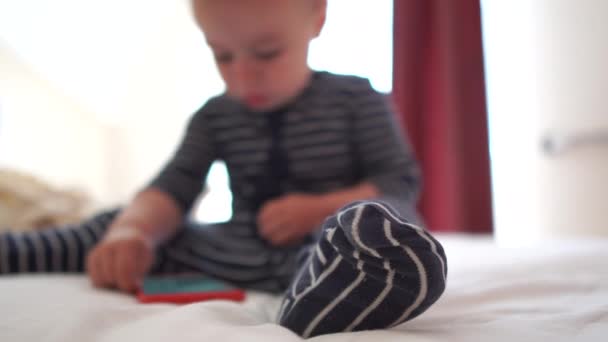 4k - 一个可爱的婴儿点击智能手机，坐在大床上 — 图库视频影像