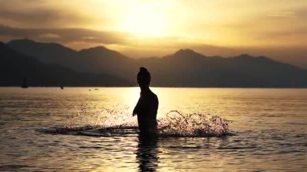 Siilhouette dívky, která v pomalém pohybu při východu slunce rozstříkne vodu v moři — Stock video