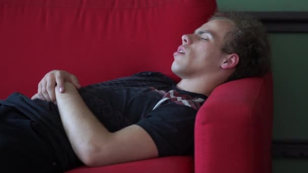 Giovane uomo biondo sdraiato e dormire su un divano rosso in uno studio bianco in slo-mo — Video Stock