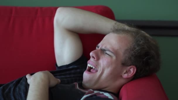 Молодой комик-блондин лежит и зевает на красном диване в студии в сло-мо — стоковое видео