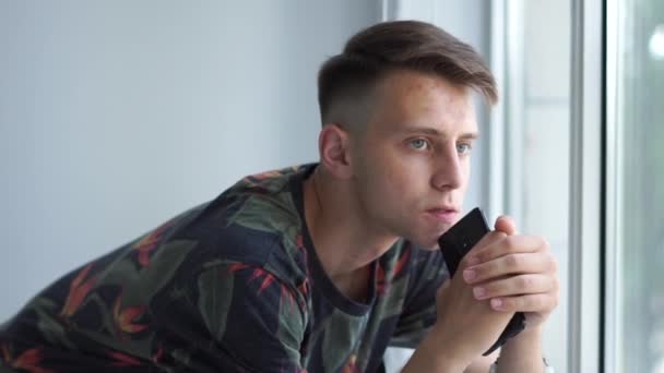 Mladý muž, který si udržuje telefon a dívá se z okna ve studiu v slo-mo — Stock video