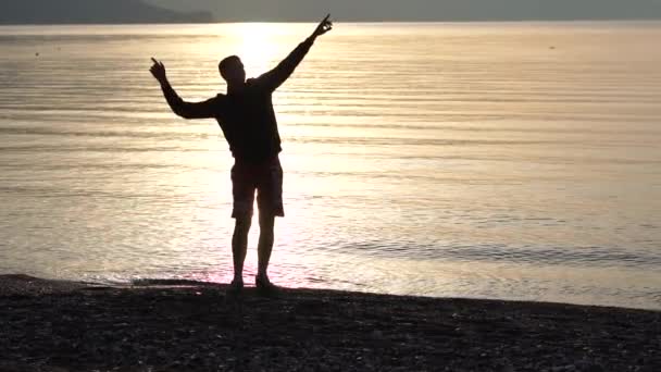 Молодой человек танцует на берегу моря на восходе солнца в замедленной съемке — стоковое видео