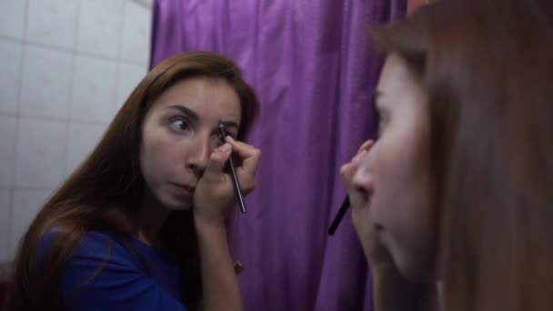 Elegante chica de cabello castaño poniendo maquillaje en su cara ante un espejo en una habitación — Vídeos de Stock
