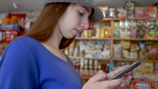 Chica de pelo castaño inteligente navegando por la red en su teléfono en una pequeña tienda en slo-mo — Vídeos de Stock