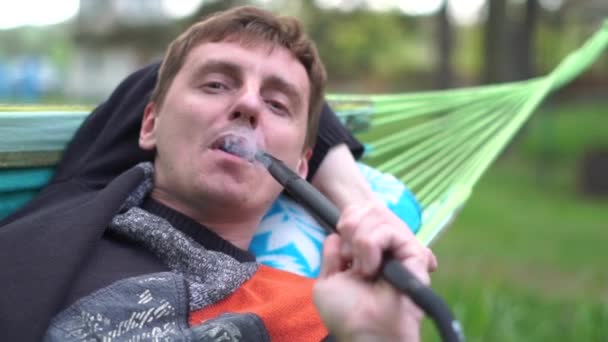 Boldog szőke férfi feküdt egy nád függőágyban dohányzás vízipipát nyáron slo-Mo