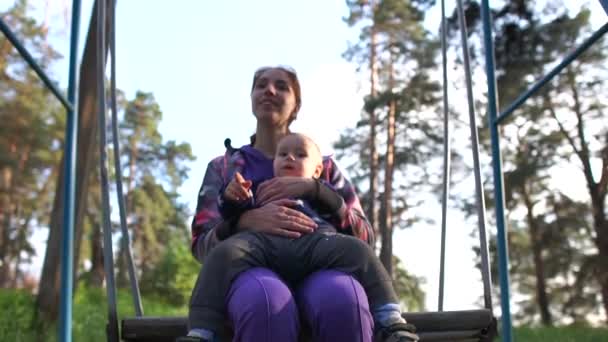 Jovem alegre com sua mãe sorridente balançando em um balanço metálico na primavera em slo-mo — Vídeo de Stock