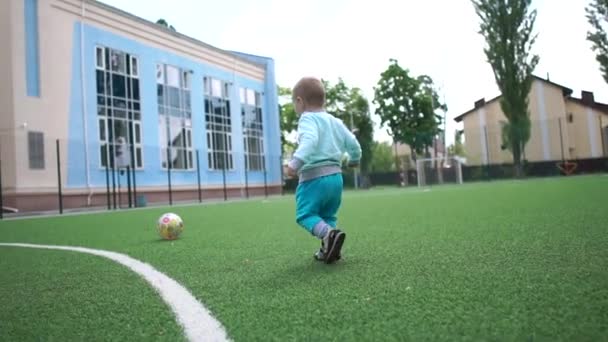 Chico rubio alegre corriendo en un campo de fútbol a una gran bola en primavera en slo-mo — Vídeos de Stock