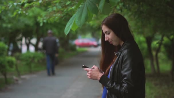 Atrakcyjna dziewczyna siedzi i przeglądania sieci na jej telefon na zewnątrz wiosną w SLO-mo — Wideo stockowe