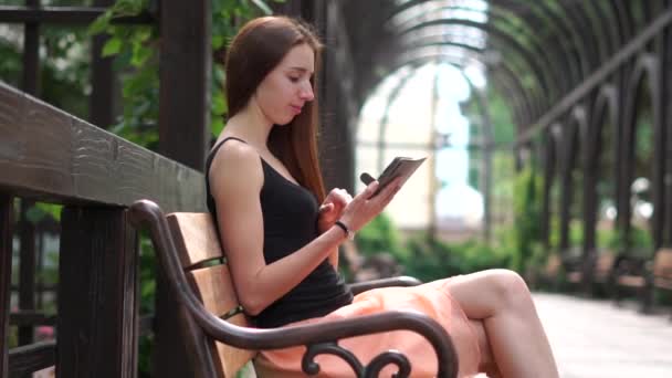 Chica se sienta en un banco e imprime un mensaje en un teléfono inteligente en el parque, cámara lenta — Vídeo de stock