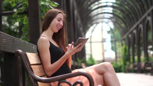 Chica lee alegremente algo en un teléfono inteligente, sentado en un parque en un banco — Vídeos de Stock