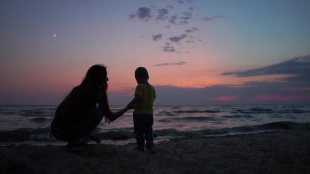 Neşeli anne çocuğuyla birlikte deniz kıyısında oturuyor. Yavaş çekimde muhteşem gün batımında. — Stok video