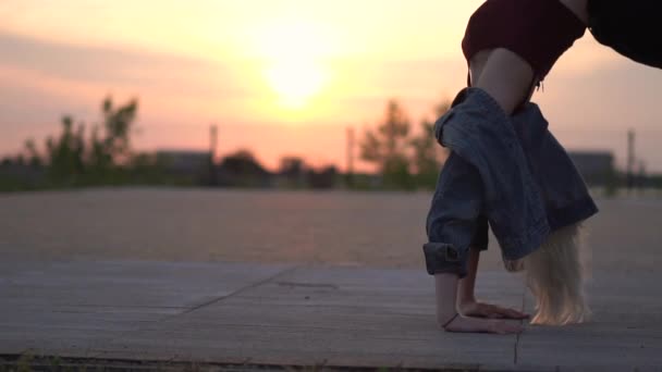 Giovane e flessibile ragazza in pantaloncini e una giacca di jeans fa un ponte al tramonto, l'azione al rallentatore. — Video Stock