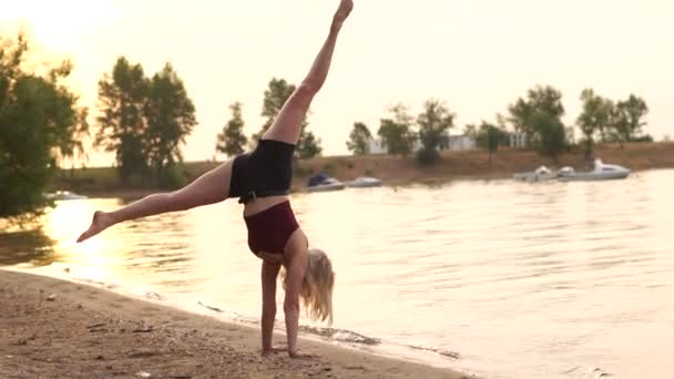 Jovem faz exercícios de ginástica ao pôr do sol na margem do lago em câmera lenta. — Vídeo de Stock