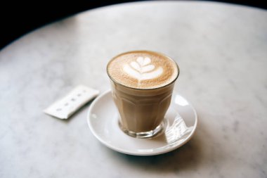 Beyaz yuvarlak mermer masanın üzerinde latte sanat ile bir fincan kahve. Şeker kesesi fincanın yanında..