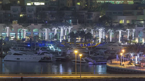 Water canal and promenade on Dubai Marina skyline at night timelapse. Residential towers with lighting and illumination. Floating yachts and boats with traffic on a bridge near skyscrapers