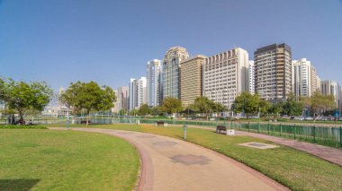 Abu Dabi 'nin sahil şeridi boyunca uzanan Corniche Bulvarı plajı parkında arka planda gökdelenler var. Lake Park 'ın manzarası. Bir tarafta ağaçlar. Güneşli bir günde mavi gökyüzü