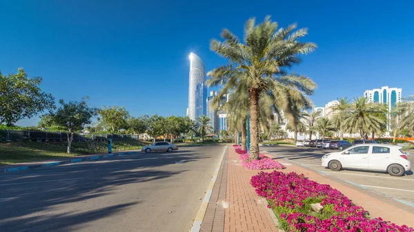Corniche boulevard beach park along the coastline in Abu Dhabi timelapse hyperlapse with skyscrapers on background. View from family park. Palms and flowers on a side. Blue sky at sunny day