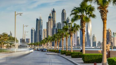 Palm Jumeirah rıhtımında yol kenarında palmiyeler ve gün batımından önce trafik zamanı. Dubai, Birleşik Arap Emirlikleri. Arka planda Dubai marina kuleleri