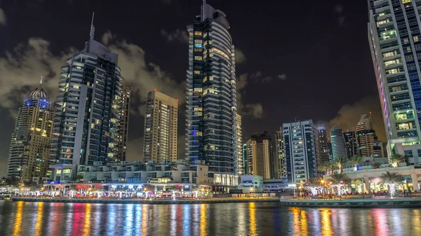 Vew of Dubai Marina promenade with yachts and modern Towers reflected in water from embankment in Dubai night timelapse hyperlapse, United Arab Emirates. Dubai Marina is a district in Dubai and an artificial canal city.