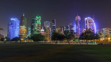 Geceleri Doha 'nın ufuk çizgisi. Yıldızlı gökyüzü Park Timelapse hiper hızından görünüyor, Katar. Ön planda ağaçlar. Aydınlatılmış gökdelenler ve kuleler