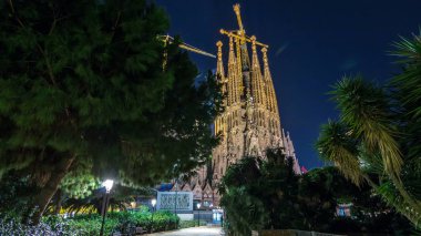 Top of Illuminated Sagrada Familia, a large Roman Catholic church in Barcelona, Spain night timelapse hyperlapse. Spires and cranes. View from park clipart