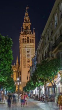 Barrio Santa Cruz 'un Seville, İspanya' daki gece zamanı. Arka planda Giralda kulesi aydınlanıyor. Turistler ve yerliler kafeler ve dükkanlarla dolu büyüleyici kaldırım taşları caddelerinde yürüyorlar..