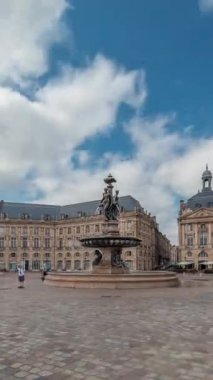 Place de la Bourse zaman atlaması Fransa 'nın Bordeaux şehrinde, Bulutlu mavi gökyüzünün altında klasik mimari ile çevrili Üç Zarafet Çeşmesi ile birlikte. Meydanda yürüyen insanlar.