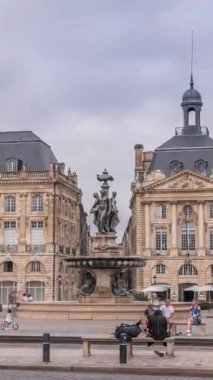 Place de la Bourse zaman atlaması Fransa 'nın Bordeaux şehrinde, Bulutlu mavi gökyüzünün altında klasik mimari ile çevrili Üç Zarafet Çeşmesi ile birlikte. İnsanlar ana meydanda bir tramvay durağında