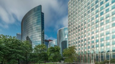 Panorama modern ofis binalarının zaman çizelgesini gösteriyor. Parktaki yeşil ağaçlar tarafından çerçevelenmiş parlak mavi gökyüzü üzerinde yansımaları ve röfleleri olan cam gökdelenler. Brüksel 'de güneş ışığı ve geçen bulutlar