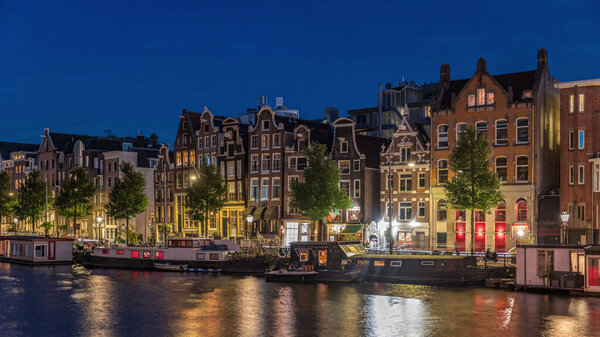Panoramic timelapse hyperlapse of the illuminated famous Dancing Houses along the Amstel River day to night transition. Water channels with houseboats. Iconic landmark in Amsterdam, Netherlands