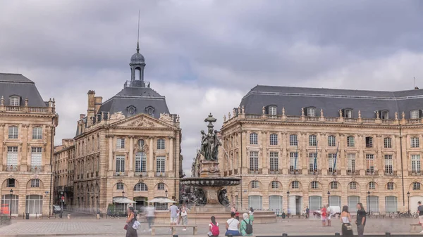 Place de la Bourse zaman atlaması Fransa 'nın Bordeaux şehrinde, Bulutlu mavi gökyüzünün altında klasik mimari ile çevrili Üç Zarafet Çeşmesi ile birlikte. İnsanlar ana meydanda bir tramvay durağında