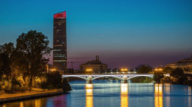 Guadalquivir Nehri yakınlarındaki Torre Sevilla gökdeleninin yer aldığı Seville 'in ufuk çizgisi gece gündüz geçiş zamanı. Aydınlatılmış Triana Köprüsü su, modern ve geleneksel mimariyi yansıtıyor