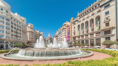 Panorama, İspanya 'nın Valencia şehrinde Belediye Meydanı' nda (Plaza del Ayuntamiento Meydanı) fıskiyeyi gösteriyor. Büyük mimari, çiçek açan yeşillik ve mavi gökyüzünün altında su jetleri. İnsanlar meydanda geziniyor.