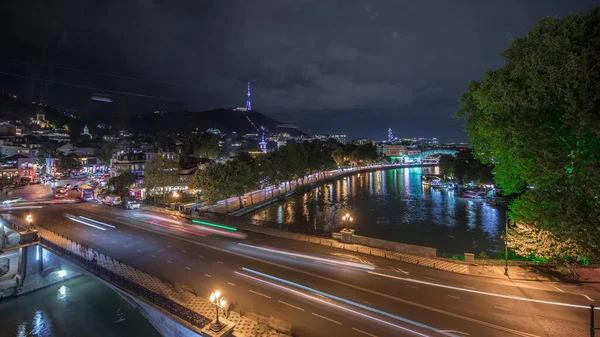 Gürcistan 'ın Tiflis şehrinde ahşap oyulmuş balkonlu renkli geleneksel evleri gösteren panorama. Aydınlatılmış binalar ve nehir üzerindeki bir köprüde trafik