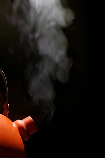 Orange kettle with boiling water and steam escaping from the spout against black background.