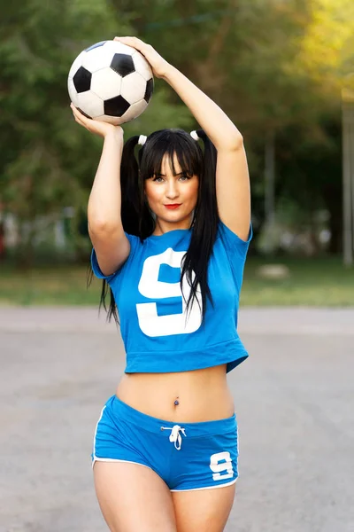 Sports girl in blue sports uniform posing with a ball as a football player against blurred background.