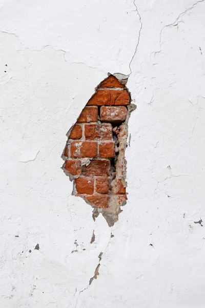 Old cracked white wall with broken plaster through which red bricks are visible