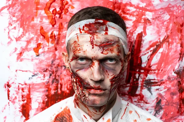 Creative art make-up for Halloween night, man face painted with cosmetics, wounds flowing blood slapping white shirt. Scary zombie. Bandage on his head, turning into a terrible nightmare horror party
