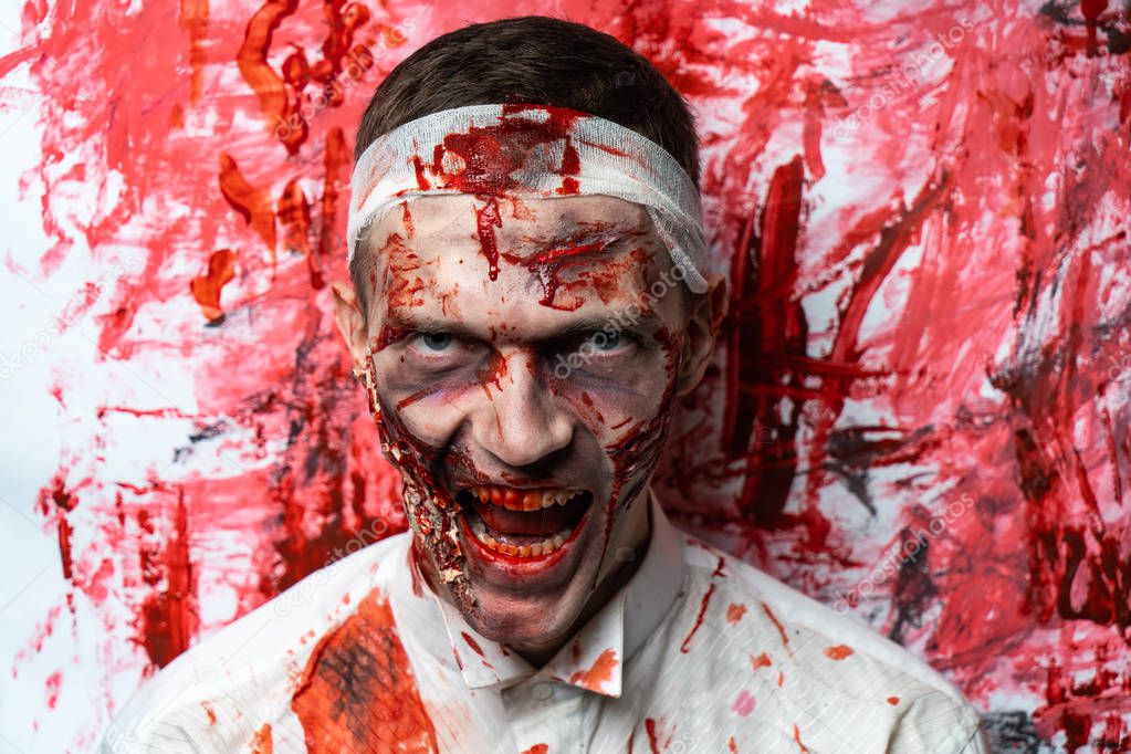 Creative art make-up for Halloween night, man face painted with cosmetics, wounds flowing blood slapping white shirt. Scary zombie. Bandage on his head, turning into a terrible nightmare horror party