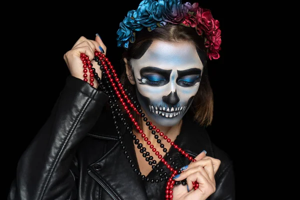 Creative makeup for Halloween party. Voodoo girl with picture makeup on her face, spooky scary skull. Holds beads jewelry. White teeth, black eyebrows drawn. Wreath of flowers, cool leather costume