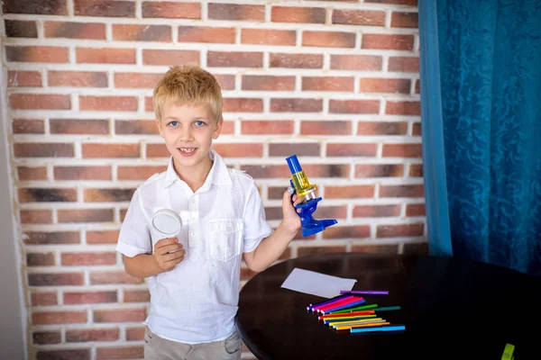 Little boy working with microscope for studying microbes. Concept love of science, thirst for knowledge and discovery. Blond hair handsome face, white clean shirt preparing for school. Red bricks wall