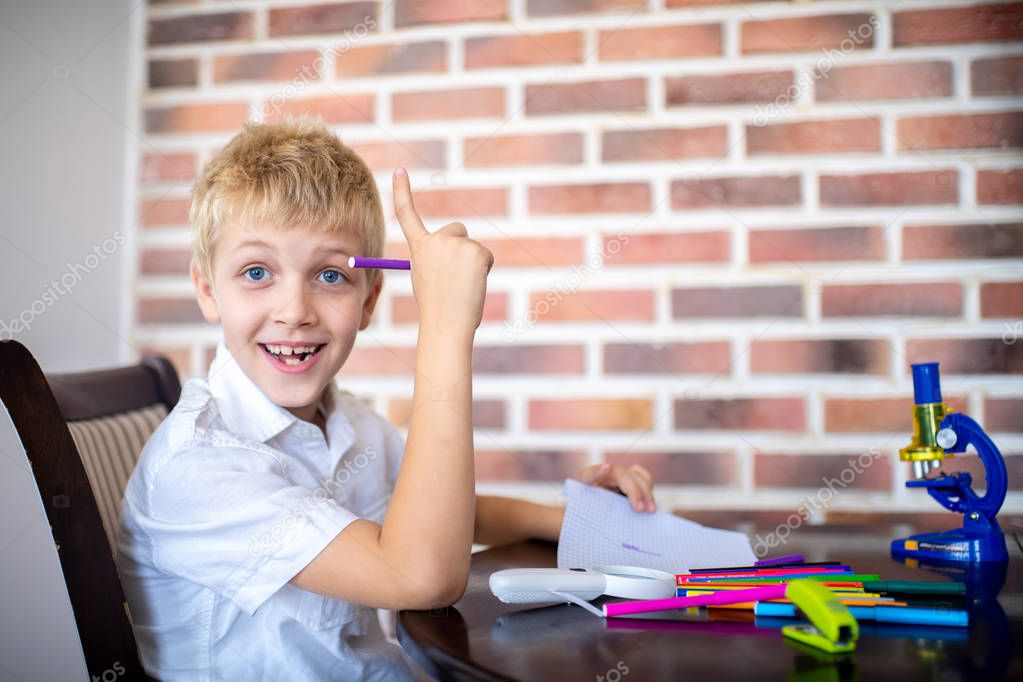 Happy cheerful boy came up with an idea. Young pupil student is sitting in a room near a brick wall, on the table are scattered markers and office supplies. Educational system lifestyle of new people