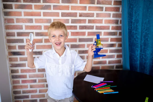 Little boy working with microscope for studying microbes. Concept love of science, thirst for knowledge and discovery. Blond hair handsome face, white clean shirt preparing for school. Red bricks wall