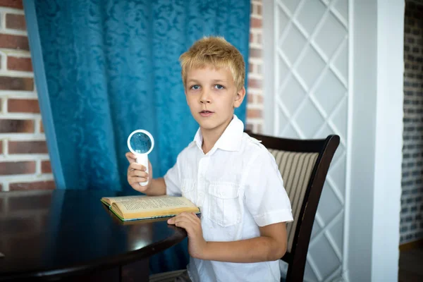 Young handsome boy, white hair, an official short-sleeved shirt. He is sitting at the table holding a magnifying glass. Attentive reading of the book, game of detectives, true love of education school