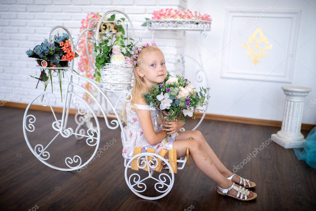 Little beautiful girl sits on a cart near a giant metal basket with colorful flowers. Blonde hair decorated with wreath. Big gift for beloved mother, conceptual photo of caring, support in the family